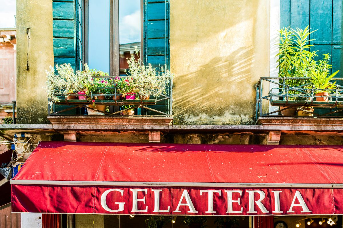 Fargerik fasade med rød markise og teksten "Gelateria" – en klassisk italiensk iskrembutikk med blomster på balkongen over.