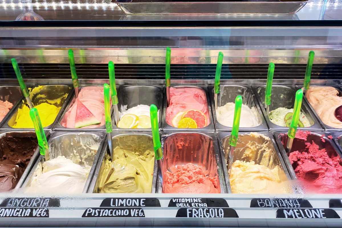 Fargerik utstilling av italiensk gelato i ulike smaker: sitron, jordbær, pistasj, vannmelon og mer. Veganske alternativer er også tilgjengelige.