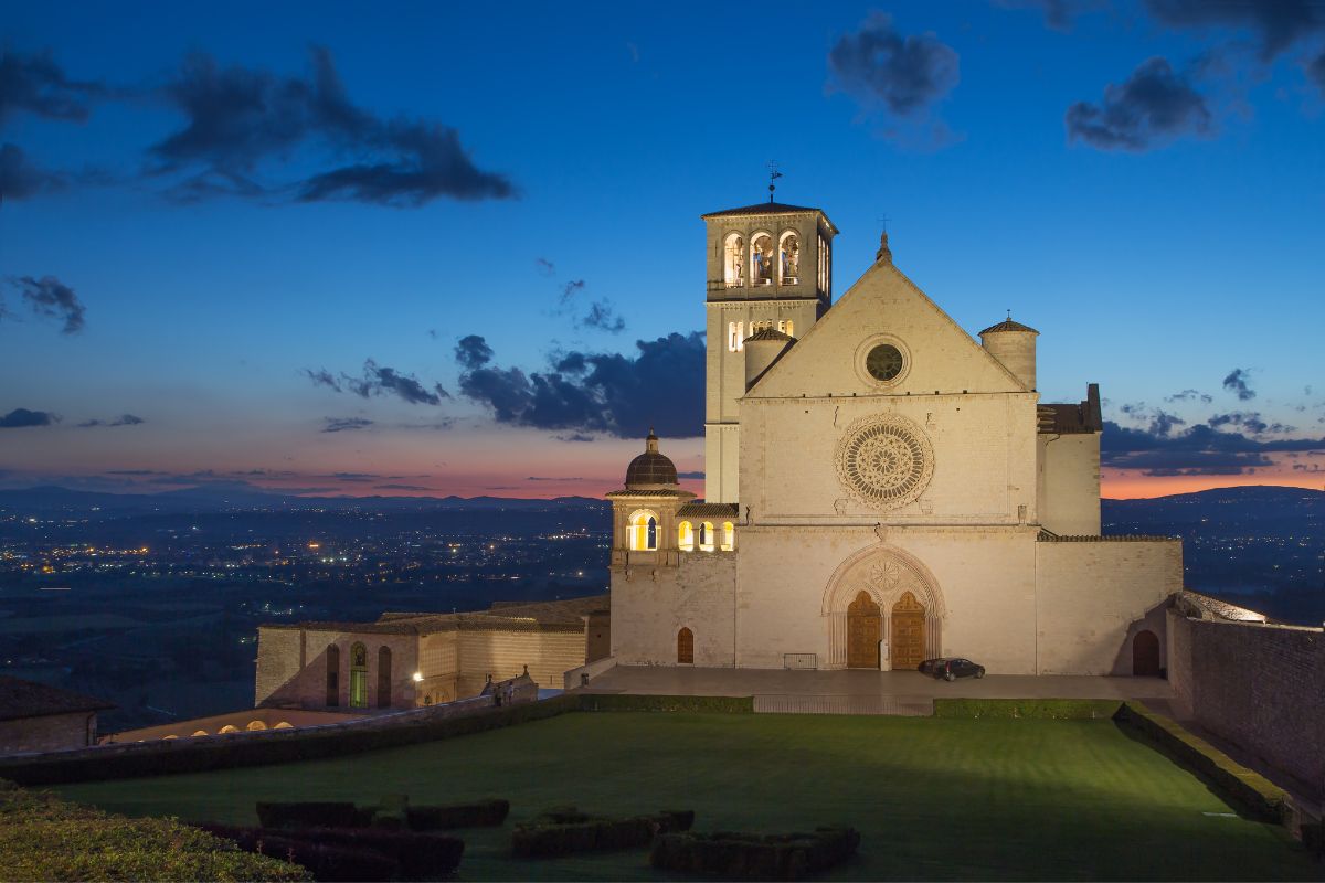 Basilica San Francesco3 Basilica di San Francesco i Assisi ved solnedgang, med fasaden og rosettvinduet opplyst mot den blå kveldshimmelen.