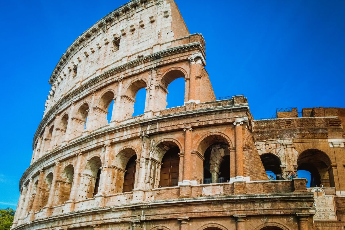 Delvis utsikt over Colosseum i Roma, med fasade av murstein og travertin under en klar, blå himmel.