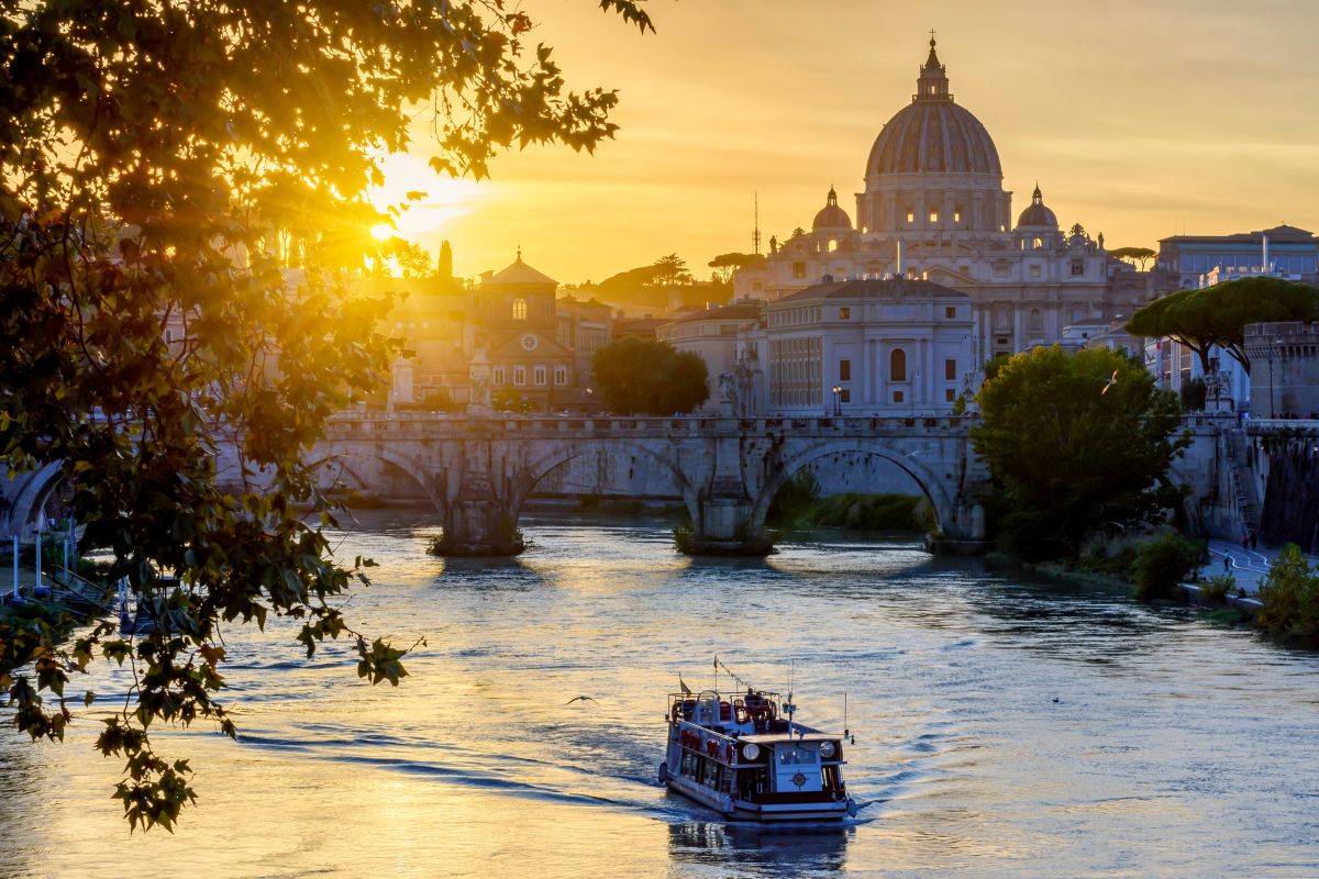 Båt som seiler på Tiberen ved solnedgang, med Peterskirken og Ponte Sant’Angelo i bakgrunnen.