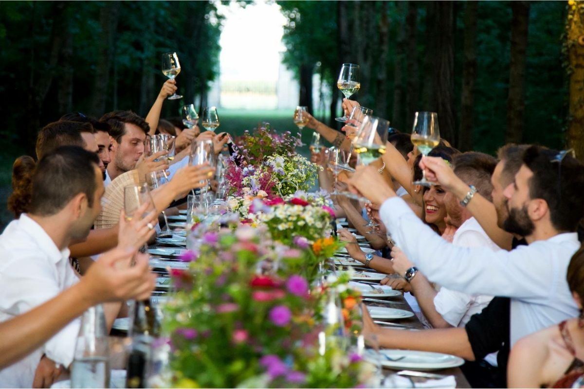 En lang utendørs festmiddag med mange mennesker som skåler med vinglass, omgitt av fargerike blomster på bordet, i en grønn allé.