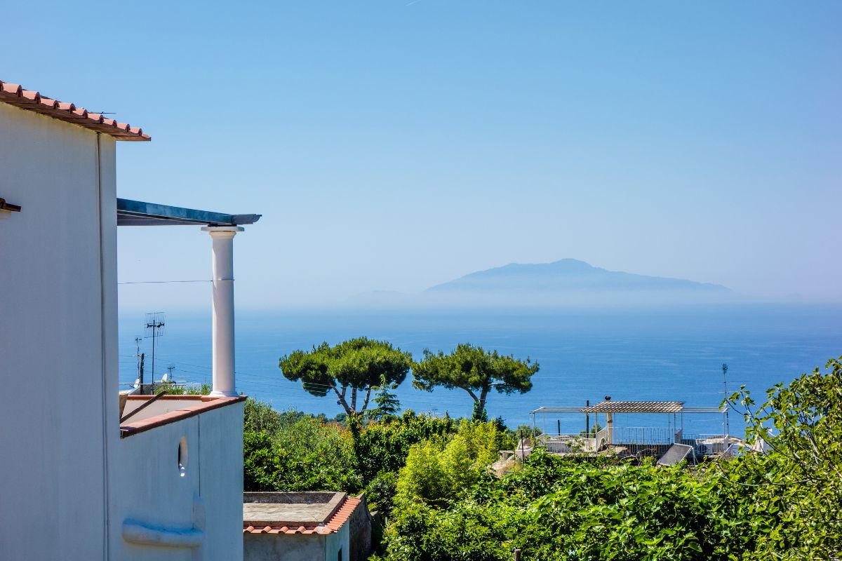 Utsikt over havet fra Ischia (Campania) med middelhavshus i forgrunnen, grønne trær og en tåkete øy i horisonten.