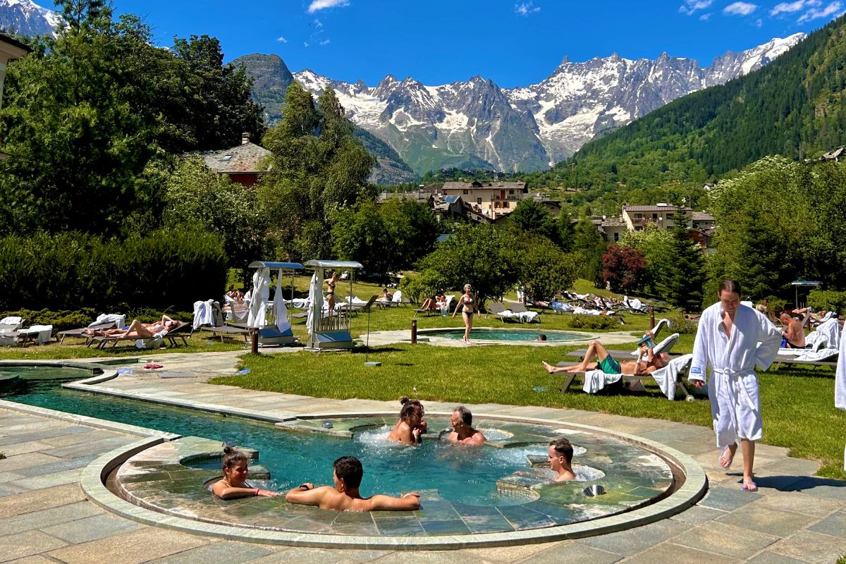 Utendørs spa-område i Pré-Saint-Didier (Valle d’Aosta) med folk som slapper av i varme bassenger og solstoler, omgitt av grønne enger og snødekte alper i bakgrunnen.