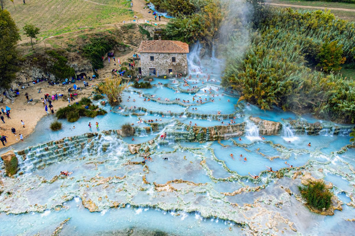 Luftfoto av de naturlige varme kildene i Saturnia, Toscana – terrasserte bassenger med blått, dampende vann omgitt av grønn vegetasjon og et gammelt steinhus. Mange mennesker bader og slapper av i vannet.