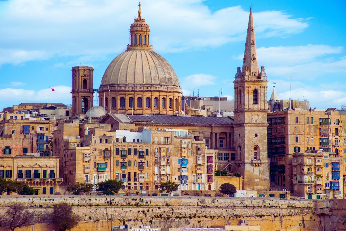 Utsikt over Valletta på Malta med den ikoniske kuppelen til Basilica of Our Lady of Mount Carmel og kirkespiret til St. Paul’s Anglican Cathedral. Byens sandfargede bygninger ligger tett ved Middelhavet under en blå himmel.