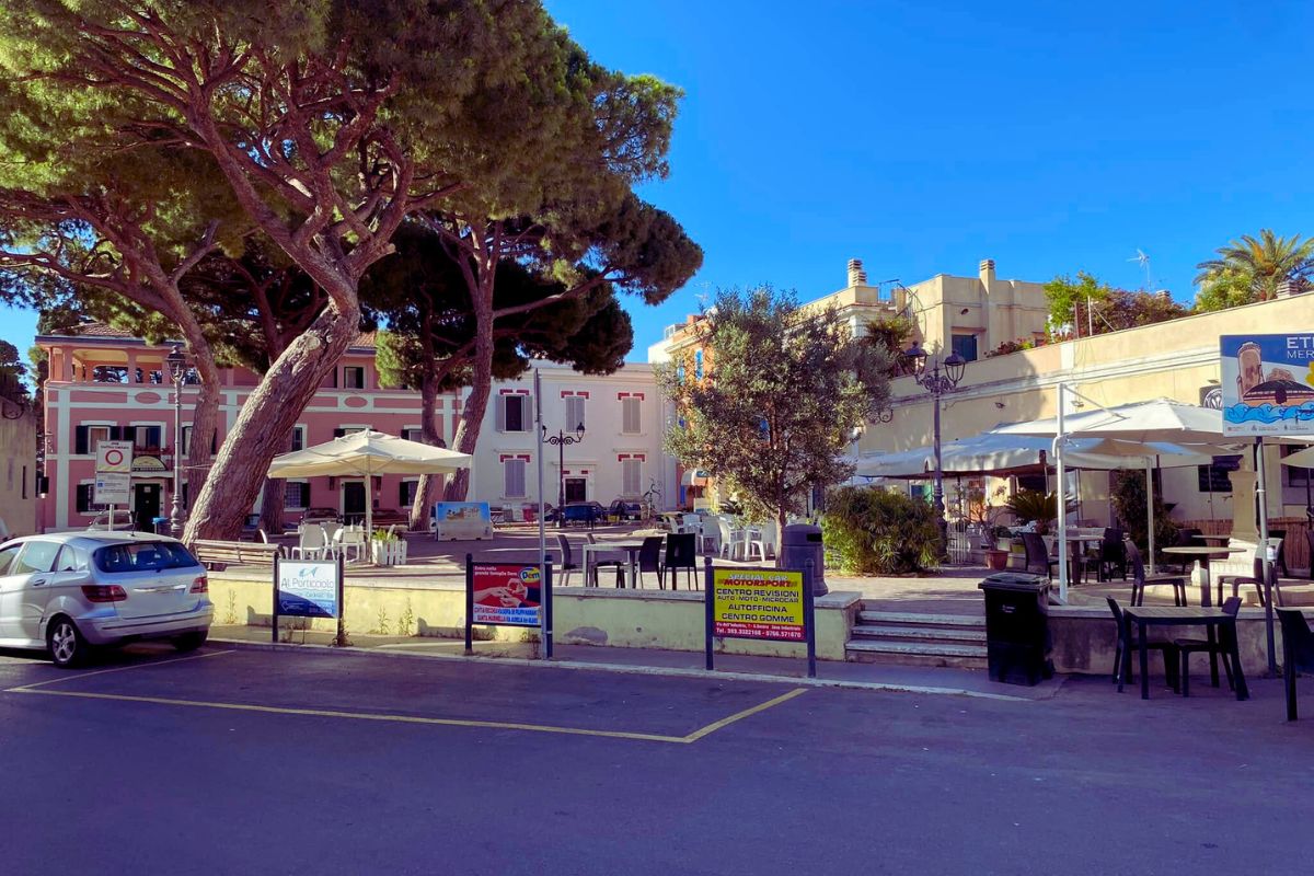 Piazza Trieste i Santa Marinella – et lite torg med kafeer, pastellfargede bygninger og store furutrær under den blå himmelen.