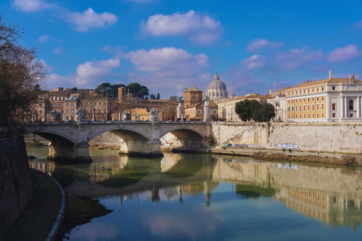 Utsikt over Roma med Ponte Sant’Angelo og Peterskirken i bakgrunnen, speilet i vannet til Tiberen under en klar blå himmel.