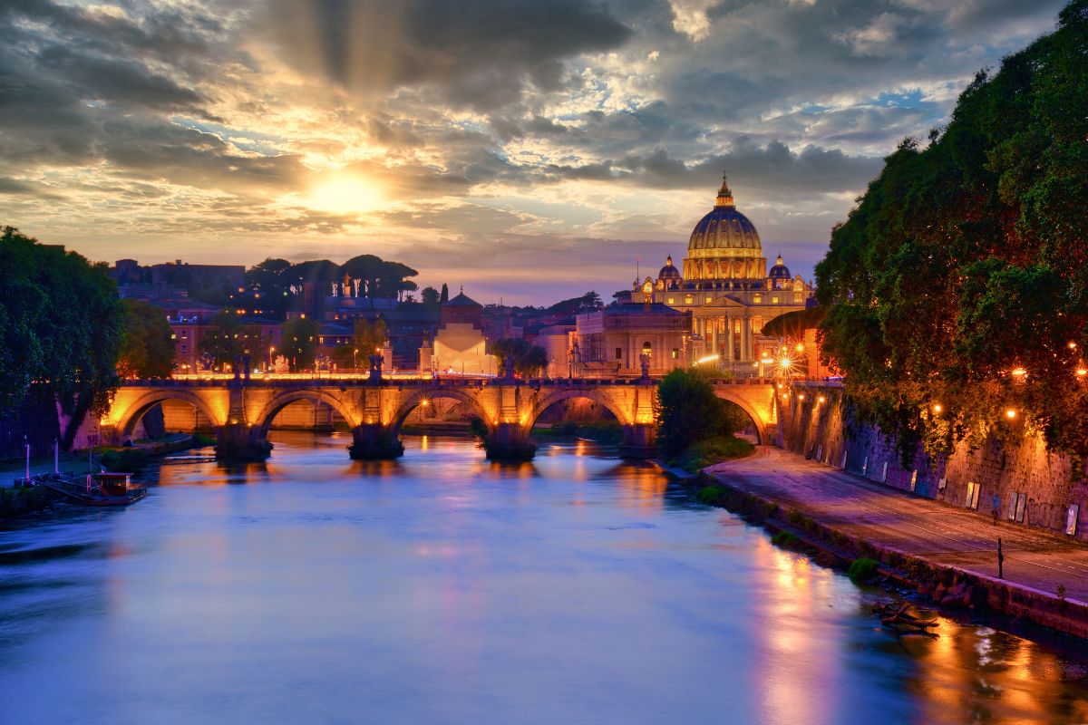 Solnedgang over Roma med Peterskirken i bakgrunnen. Den opplyste broen Ponte Sant’Angelo speiler seg i Tiber-elven, mens kveldssolen farger himmelen gyllen og dramatisk.