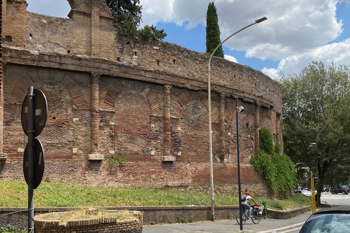 Den buede murveggen til Amfiteateret Castrense i Roma, innlemmet i Aurelianmuren, sett fra gateplan med gamle murstein, tilmurte buer og litt vegetasjon på siden.