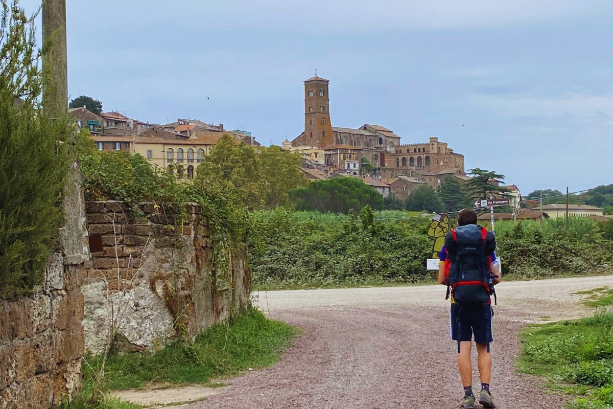 Person med ryggsekk som går langs Via Francigena mot en italiensk middelalderby med kirketårn og historiske bygninger i bakgrunnen.