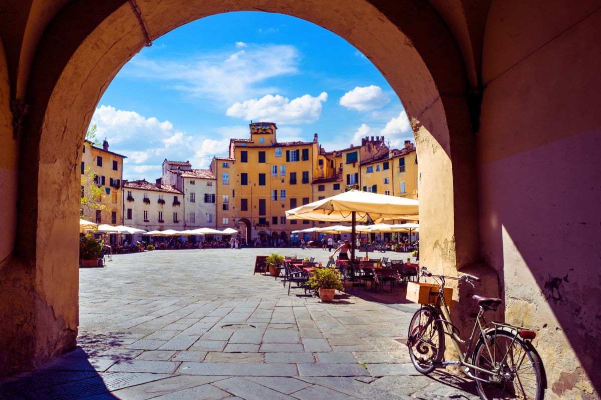 Piazza i Lucca i Toscana sett gjennom en buegang, med gule bygninger, uteservering og sykkel i forgrunnen.