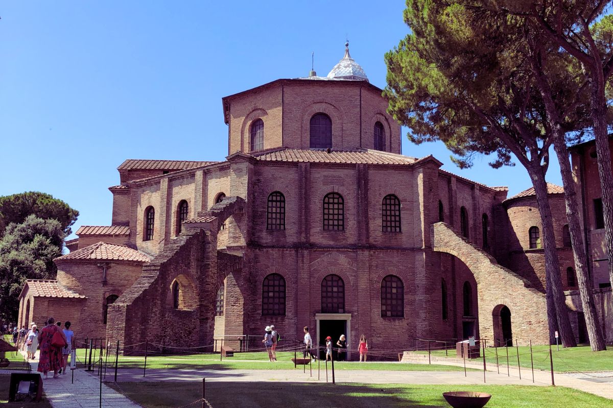 Basilikaen San Vitale i Ravenna, kjent for sin åttekantede form og bysantinske arkitektur, omgitt av trær og besøkende foran inngangen.