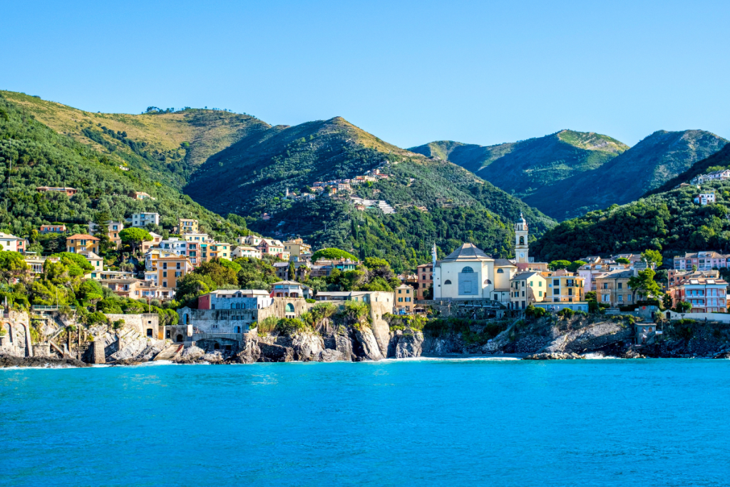 Utsikt over den liguriske kysten med fargerike hus, en kirke ved sjøen og grønne åser som stiger bratt opp fra havet. Det turkise vannet og det frodige landskapet er typisk for Liguria i Nord-Italia.