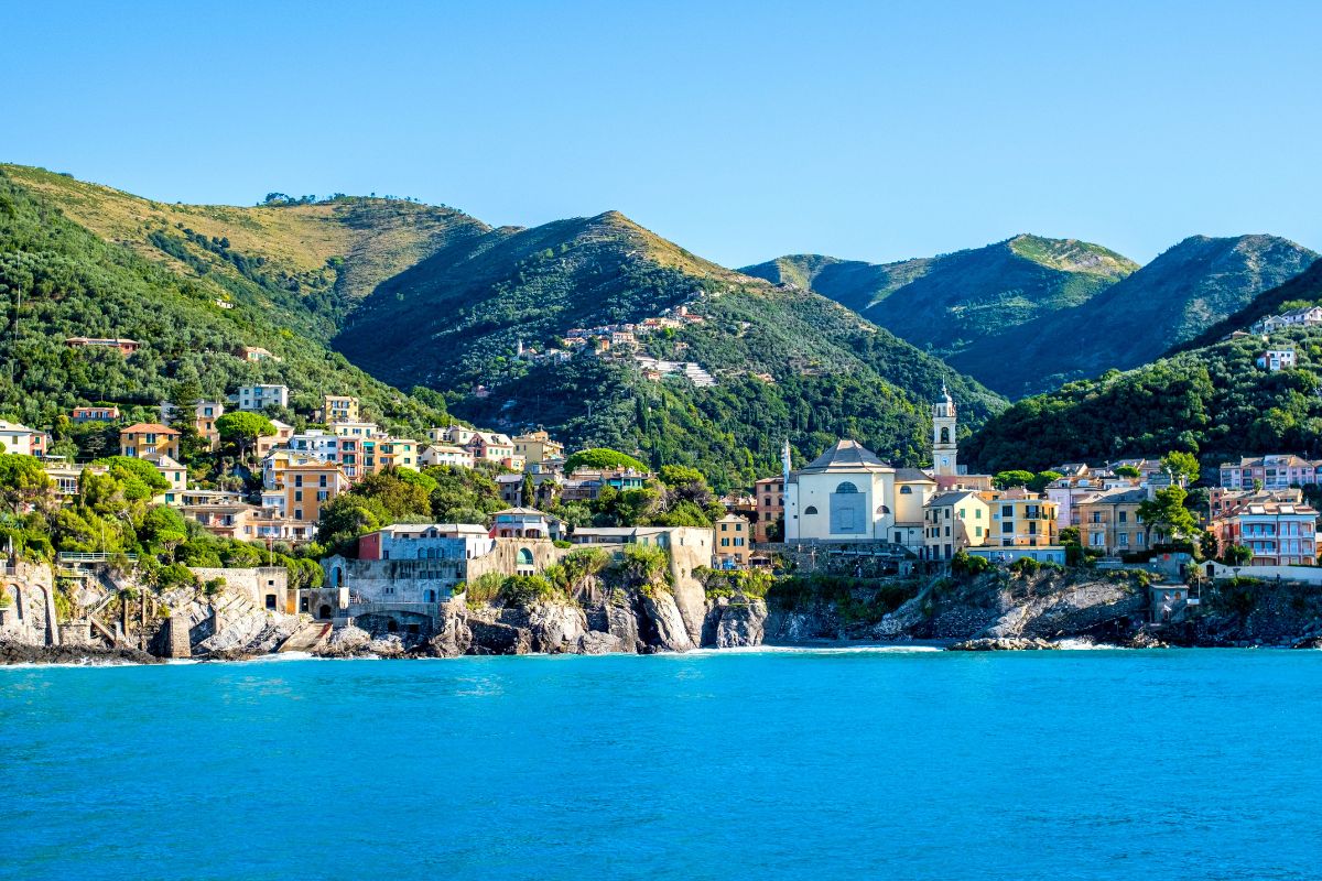 Utsikt over den liguriske kysten med fargerike hus, en kirke ved sjøen og grønne åser som stiger bratt opp fra havet. Det turkise vannet og det frodige landskapet er typisk for Liguria i Nord-Italia.