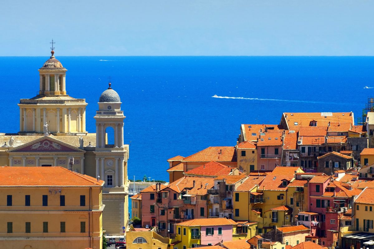 Utsikt over gamlebyen i Imperia med fargerike hus, historiske bygninger og kirketårn som reiser seg over takene. I bakgrunnen glitrer Middelhavet under en skyfri himmel, et typisk landskap fra den liguriske kysten.