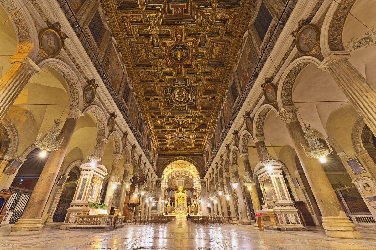 Interiøret i Santa Maria in Aracoeli i Roma. Den brede midtskipet er omgitt av antikke søyler fra romerske bygninger, mens det praktfulle forgylte taket ble laget for å feire seieren i slaget ved Lepanto i 1571. I bakgrunnen står høyalteret, omgitt av århundrer med kunst, historie og religiøse tradisjoner.